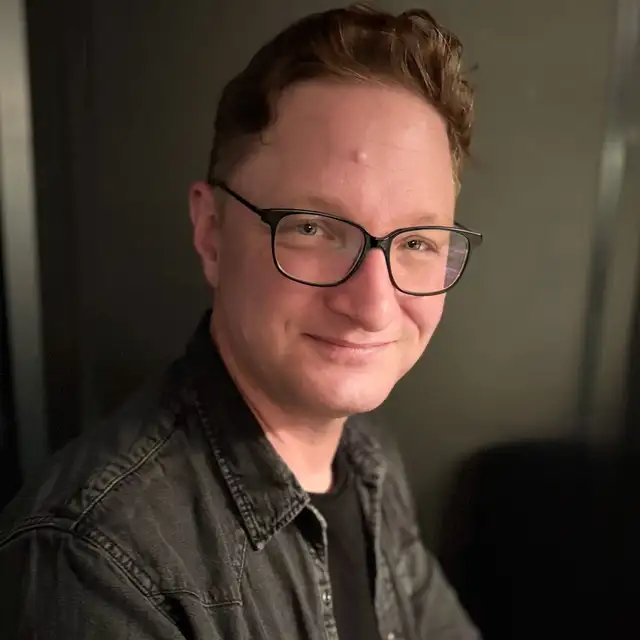 Jay Stephen Gilmore, a middle-aged man with short, wavy brown hair and blue eyes is looking directly at the camera and smiling. He is wearing rectangular black-framed glasses and a black shirt. The background appears to be a plain white wall.​​​​​​​​​​​​​​​​